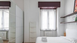 A modern hotel room with a large bed, a wardrobe, a window with curtains, and a bedside table with a lamp.