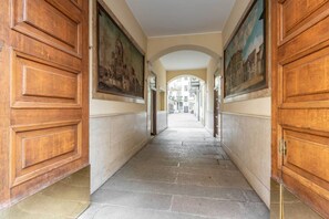 Een gang met houten deuren en schilderijen aan de wanden, die leidt naar de ingang van een gebouw.