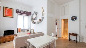 A modern living room with a white sofa, a dining table with chairs, a TV on a wooden cabinet, a large window, and a shower room visible through an open door.