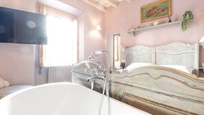 Un dormitori amb una banyera blanca gran, un panell de fusta al capçal, un quadre a la paret i una finestra amb persianes.