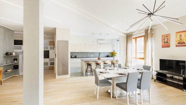 Eine moderne Küche mit weißen Schränken, einer zentralen Kochinsel und Barhocker. Es gibt einen Essbereich mit einem weißen Tisch und grauen Stühlen. Ein Fernseher ist an der Wand befestigt, und es befinden sich Bilder dort.