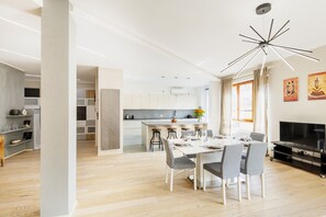 Una cocina moderna con armarios blancos, una isla central y taburetes de bar. Hay una zona de comedor con una mesa blanca y sillas grises. Un televisor está montado en la pared, y hay pinturas decorando la pared.