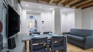 Un espace repas moderne avec une table dressée pour quatre personnes, un canapé gris et un téléviseur à écran plat fixé au mur.