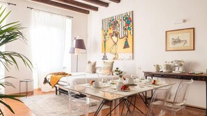 Een moderne kamer met een glazen eettafel, transparante stoelen, een bed met een witte dekbedovertrek, een lamp en een schilderij aan de muur.