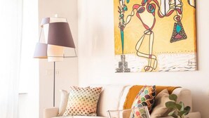 Een moderne woonkamer met een witte bank, kleurrijke kussens, een vloerlamp en een levendig abstract schilderij aan de muur.