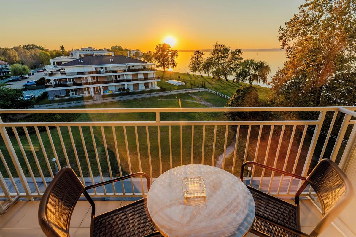 A balcony with a table and chairs, overlooking a building and a body of water during sunset.