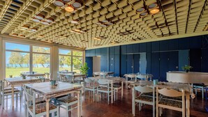 Comedor con mesas y sillas blancas, techo estampado y grandes ventanales con vistas a un parque.
