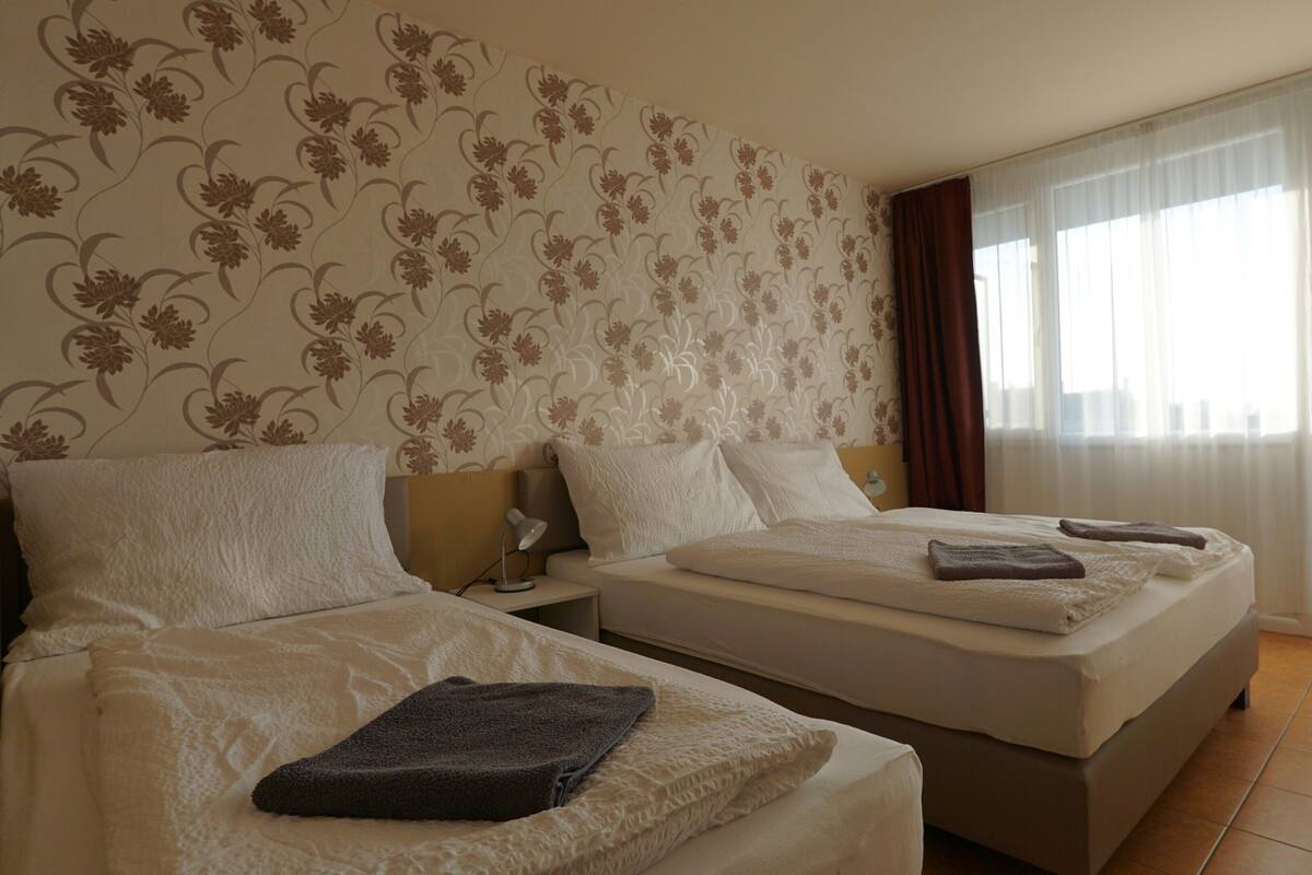 A hotel room with two beds, a nightstand, a lamp, and a large window with curtains.