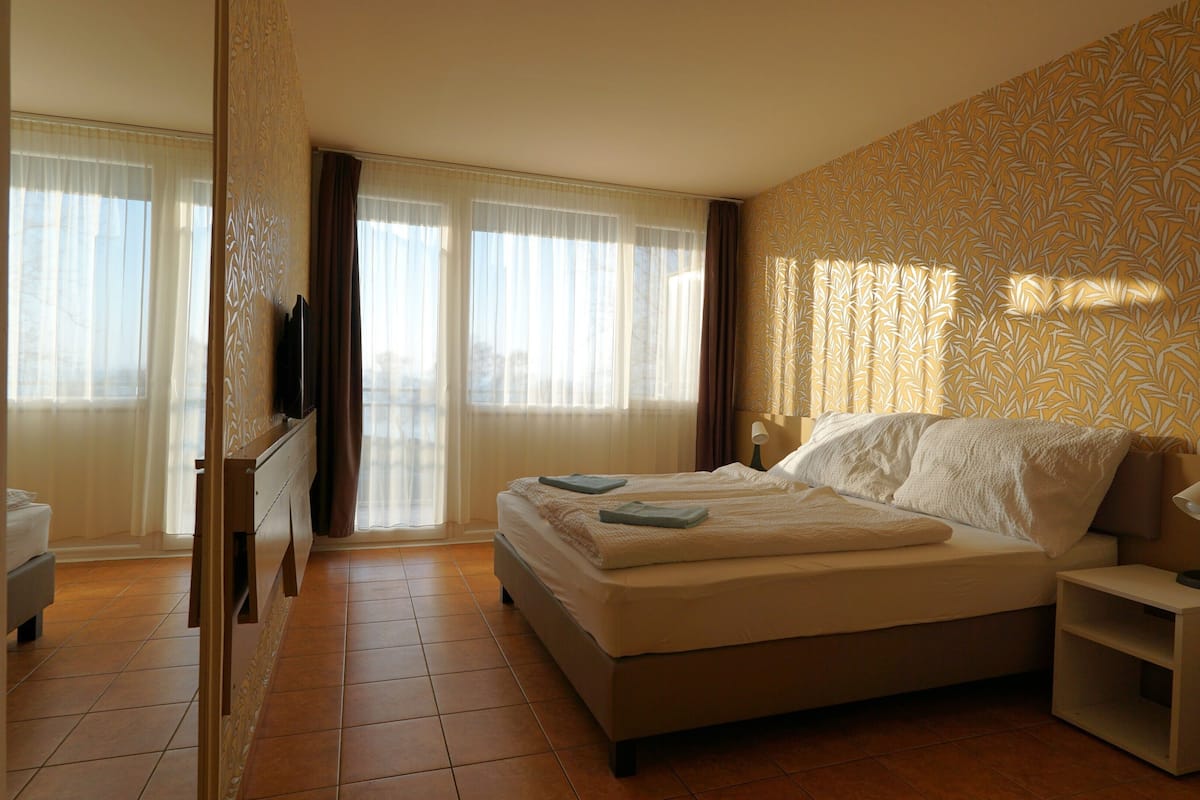 A hotel room with a bed, a TV, a bedside table, and a view of the outside through large windows.
