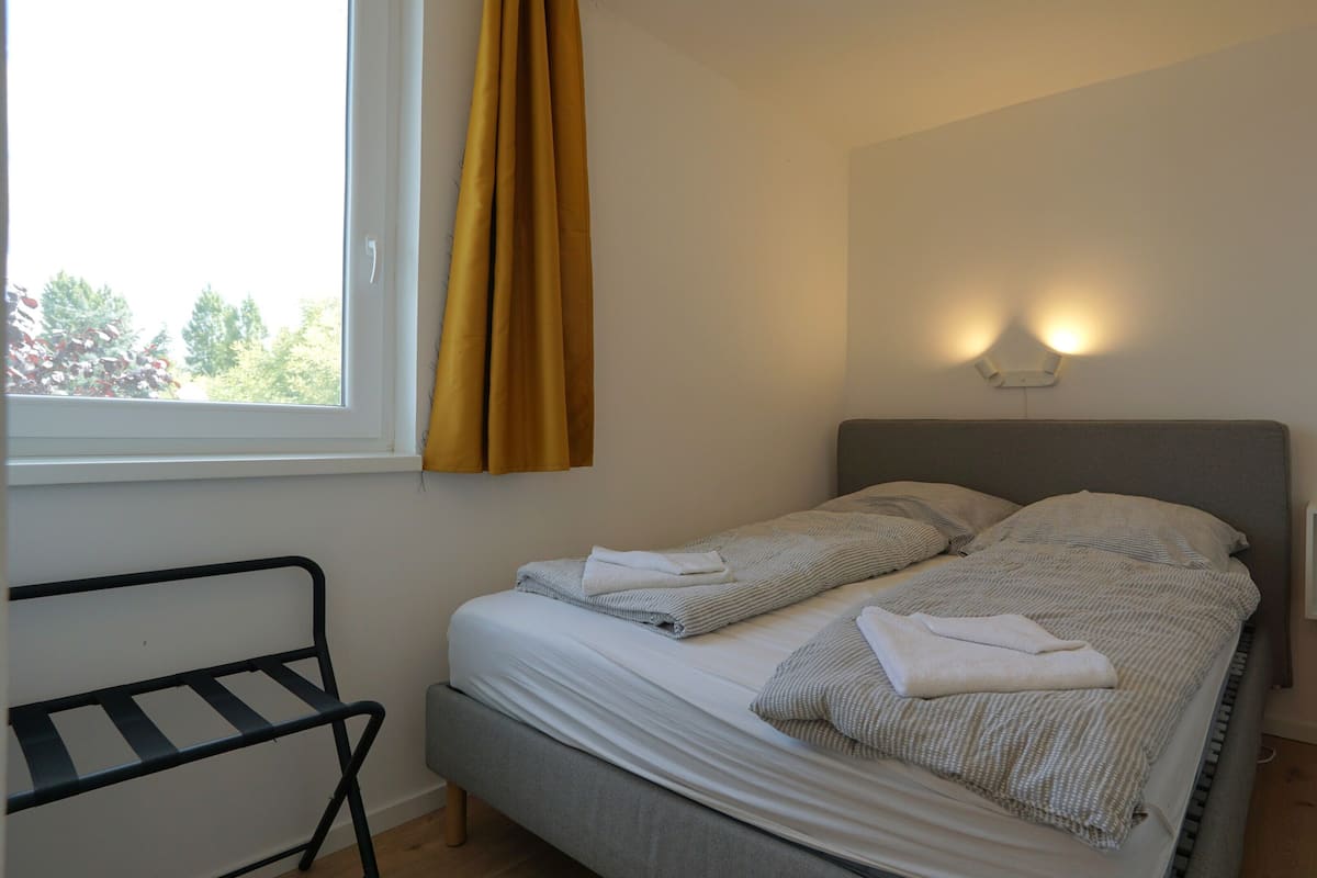A single bed with a grey headboard, a bedside table, a window with yellow curtains, and a wall-mounted light fixture.