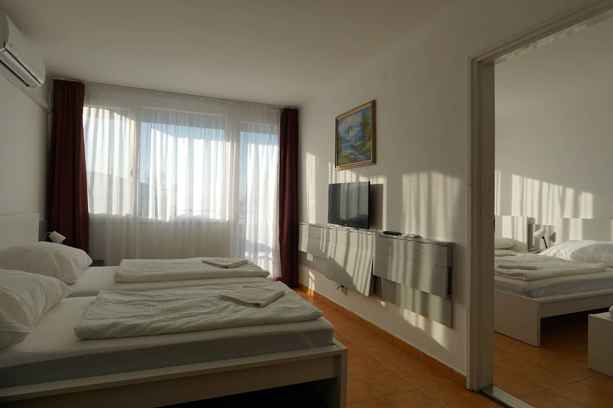 A hotel room with two beds, a TV mounted on the wall, and a large window with sheer curtains.