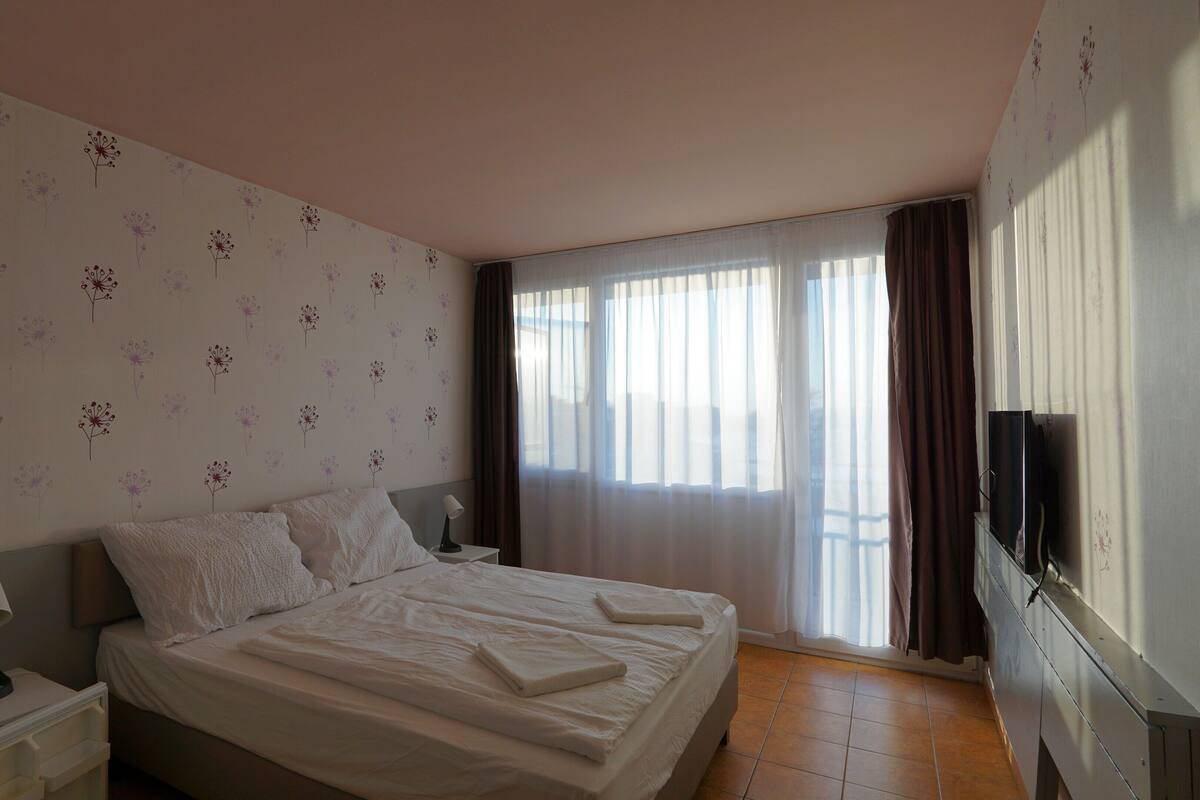 A neatly made bed with white linens, a bedside lamp, and a TV on a stand to the right.