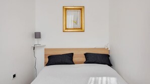 Une chambre d’hôtel au style minimaliste, avec une tête de lit en bois, un lit recouvert de linge de lit blanc, deux oreillers foncés, une lampe de chevet et un tableau encadré accroché au mur.