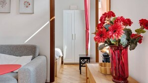 Un salon moderne avec un canapé gris, une table rouge et un vase rouge contenant des fleurs. On aperçoit une porte blanche au fond et un petit tabouret noir.