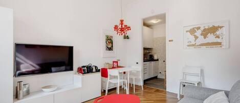 Un salon moderne avec un téléviseur à écran plat, un fauteuil rouge, un canapé gris et une cuisine en arrière-plan.