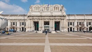 Un grand bâtiment historique doté d’une entrée centrale, d’une vaste place ouverte et de plusieurs drapeaux.