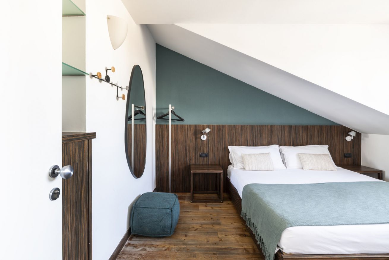 A bedroom with a sloped ceiling, a bed with white linens, a wooden floor, a small table, and a wall-mounted lamp.