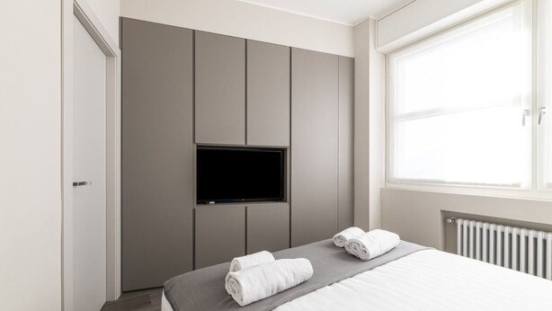 Une chambre moderne avec un lit, un téléviseur à écran plat fixé au mur et une fenêtre laissant entrer la lumière naturelle.