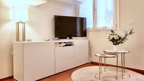 Ein moderner, gut beleuchteter Raum mit einem weißen Schrank, einem Flachbildfernseher, einem kleinen runden Tisch mit Blumenvase und einer Lampe.