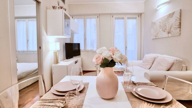 Ein modernes, helles Wohnzimmer mit Essbereich, weißen Möbeln und einem Blumenstrauß auf dem Tisch.