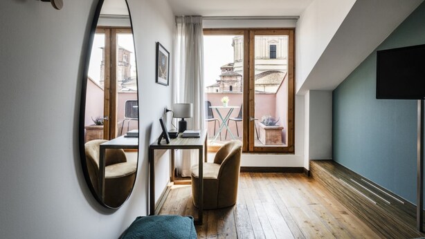 A modern room with a large mirror, a wooden floor, a desk with a chair, and a view of buildings through large windows. - Easylife - Single Suite with view (Milano)