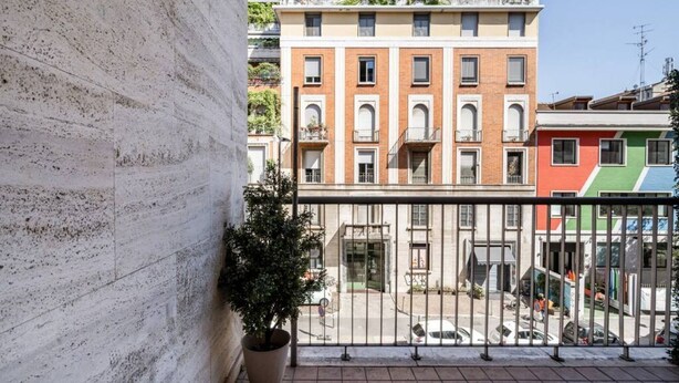 Uma varanda com um vaso de planta, um edifício com várias janelas e vista para a rua.