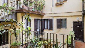 A courtyard with potted plants, a balcony with a wrought iron railing, and a building with a black door and windows with shutters. - Easylife - Comfortable studio with terrace Navigli (Milano)
