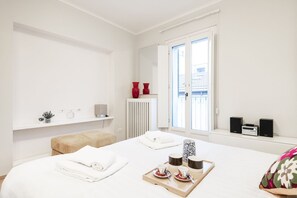 Habitación de hotel con cama, un pequeño rincón de descanso, ventana con vistas y estante con un jarrón y una radio.