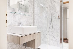 Un baño moderno con pared de mármol, repisa de vidrio, lavamanos blanco y ducha con mampara de vidrio.