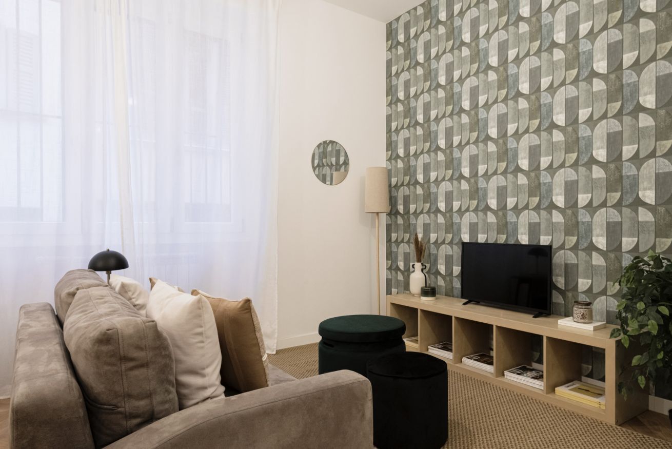 A living room with a sofa, a TV on a stand, a lamp, and a patterned wall.