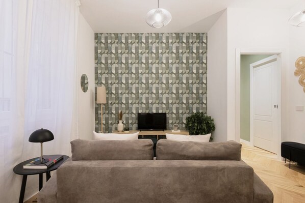 A modern living room with a grey sofa, a flat-screen TV, a small side table with a lamp, and a patterned wallpaper.