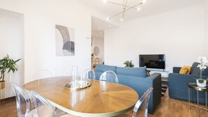 A modern living room with a round wooden dining table, clear chairs, a blue sofa, a TV, and a potted plant.
