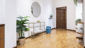A hallway with a wooden floor, a round mirror, a potted plant, a metal shelf unit, and a blue trash bin.