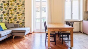 Ein modernes Wohnzimmer mit Esstisch aus Holz, grauem Sofa und ornamentalem Wandpapier.
