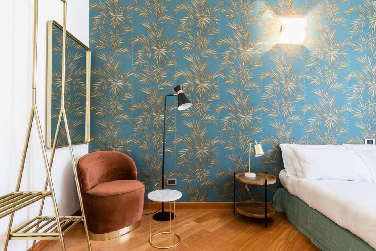 A room with a patterned blue wall, a brown chair, a small round table, a bed with white bedding, and a floor lamp.
