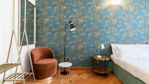 Ein Zimmer mit blauer Wand mit Muster, einem braunen Sessel, einem kleinen runden Tisch, einem Bett mit weißer Bettwäsche und einer Stehlampe.