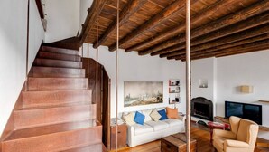 A staircase leading to a lofted area with exposed wooden beams, a white sofa with colorful cushions, a television, and a fireplace.