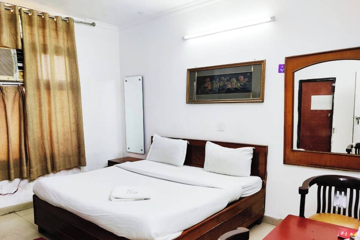 Habitación de hotel con cama de madera, una mesita, una silla, un espejo y un cuadro en la pared.
