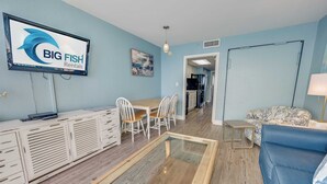 A living room with a TV displaying 'BIG FISH Rentals', a dining area with chairs, a sofa, and a glass coffee table. - Newly Updated, Beachside  Spectacular at Bay Watch (North Myrtle Beach)
