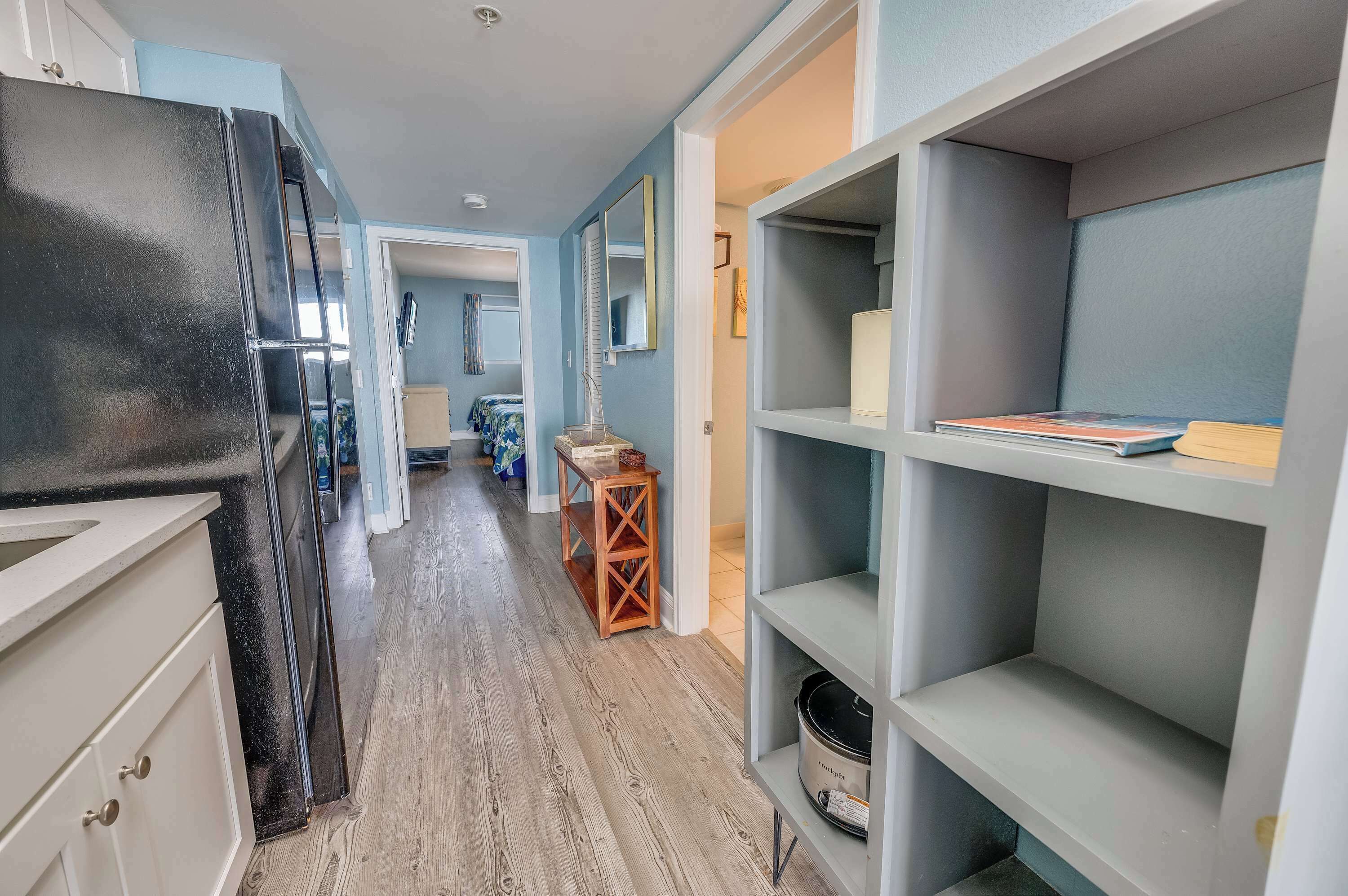A narrow hallway with a kitchen area on one side, featuring a black refrigerator and white cabinets. There are shelves with books and a small wooden table with a vase.