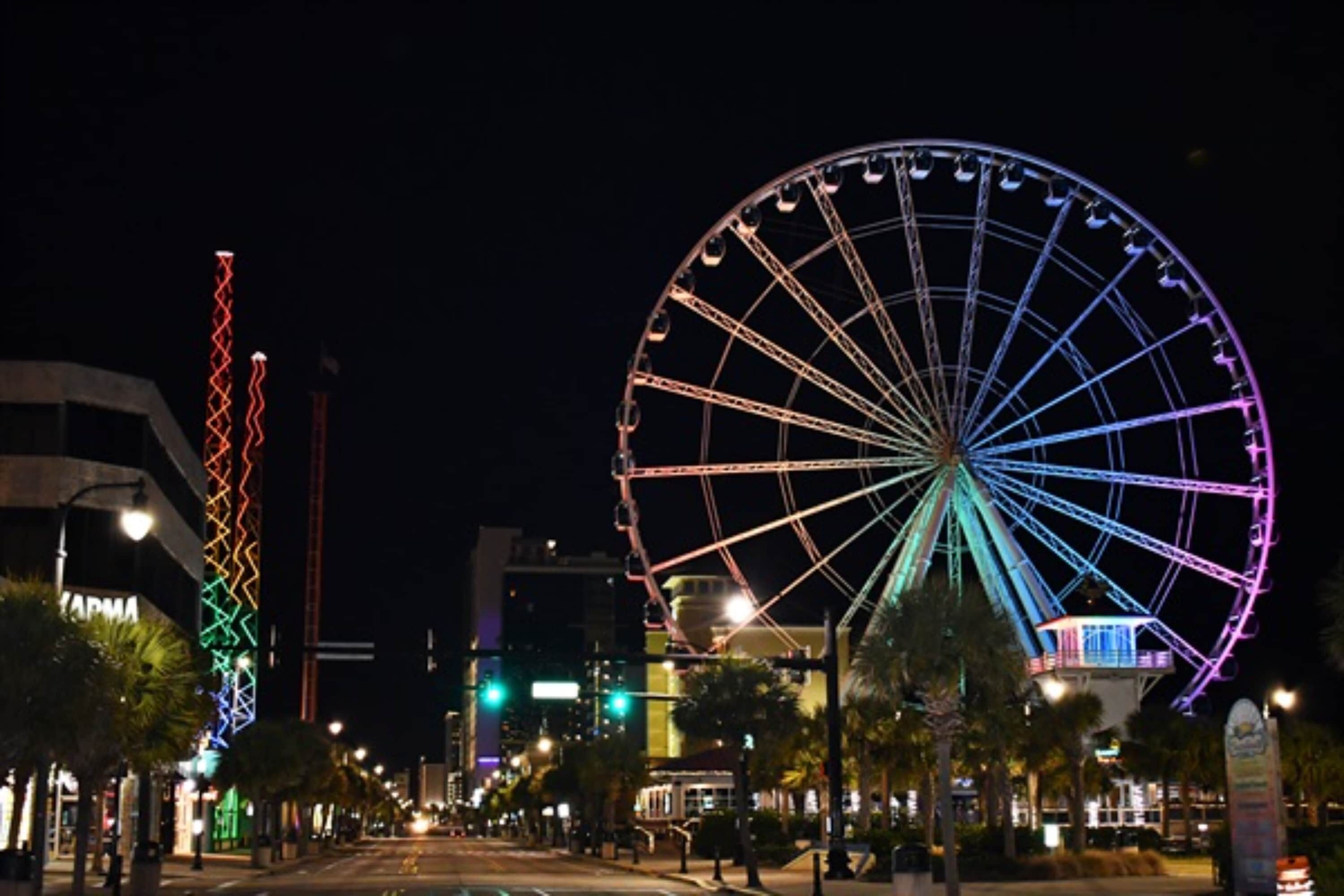 Une vue nocturne d’une rue avec une grande grande roue et un bâtiment illuminé, sur lequel on peut lire l’inscription « PARME ».