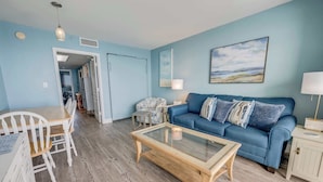 A living room with a blue sofa, a glass coffee table, and a dining area with a wooden table and chairs. - Newly Updated, Beachside  Spectacular at Bay Watch (North Myrtle Beach)