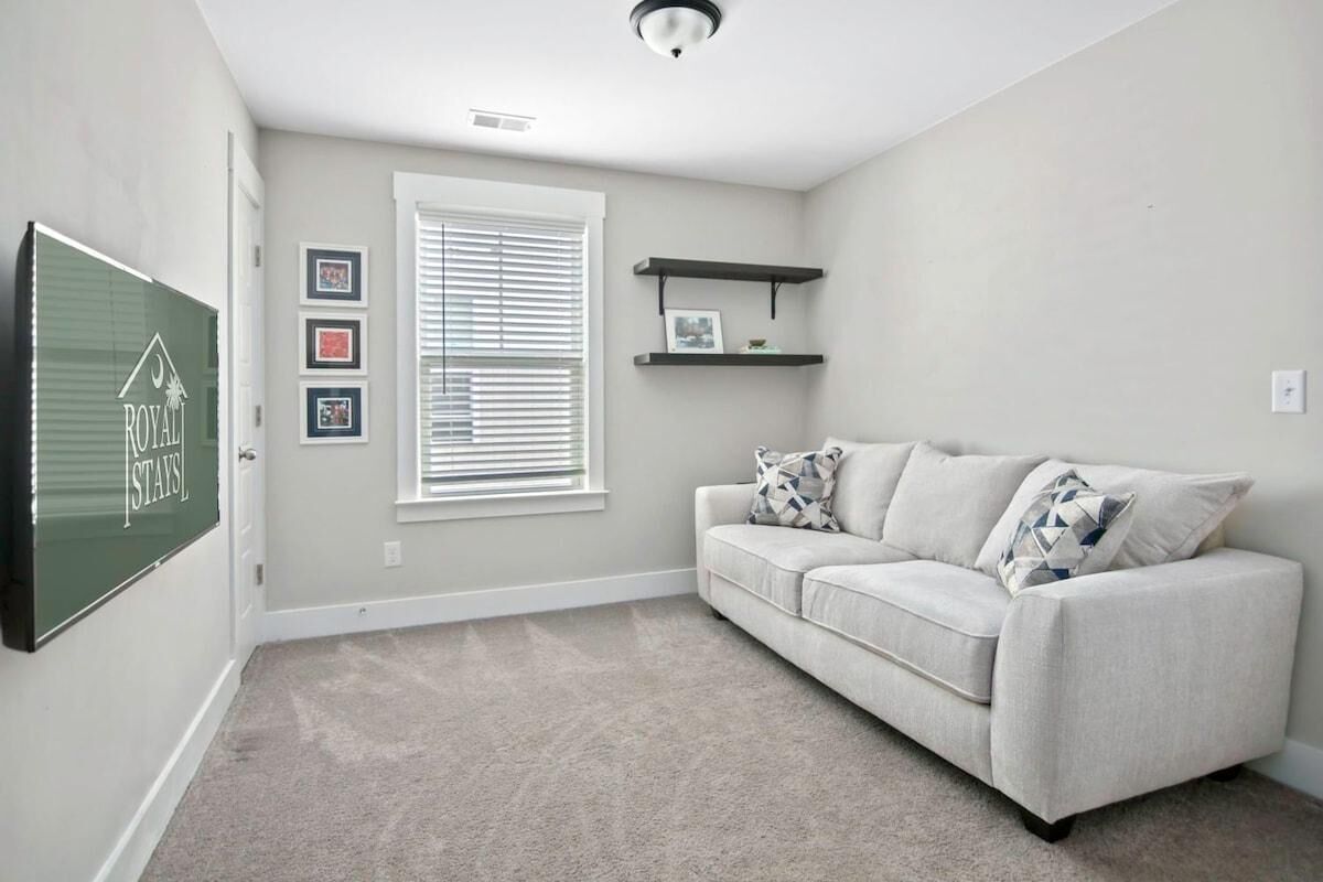A living room with a beige carpet, a light grey sofa with patterned cushions, a window with blinds, and a wall-mounted shelf with framed pictures.