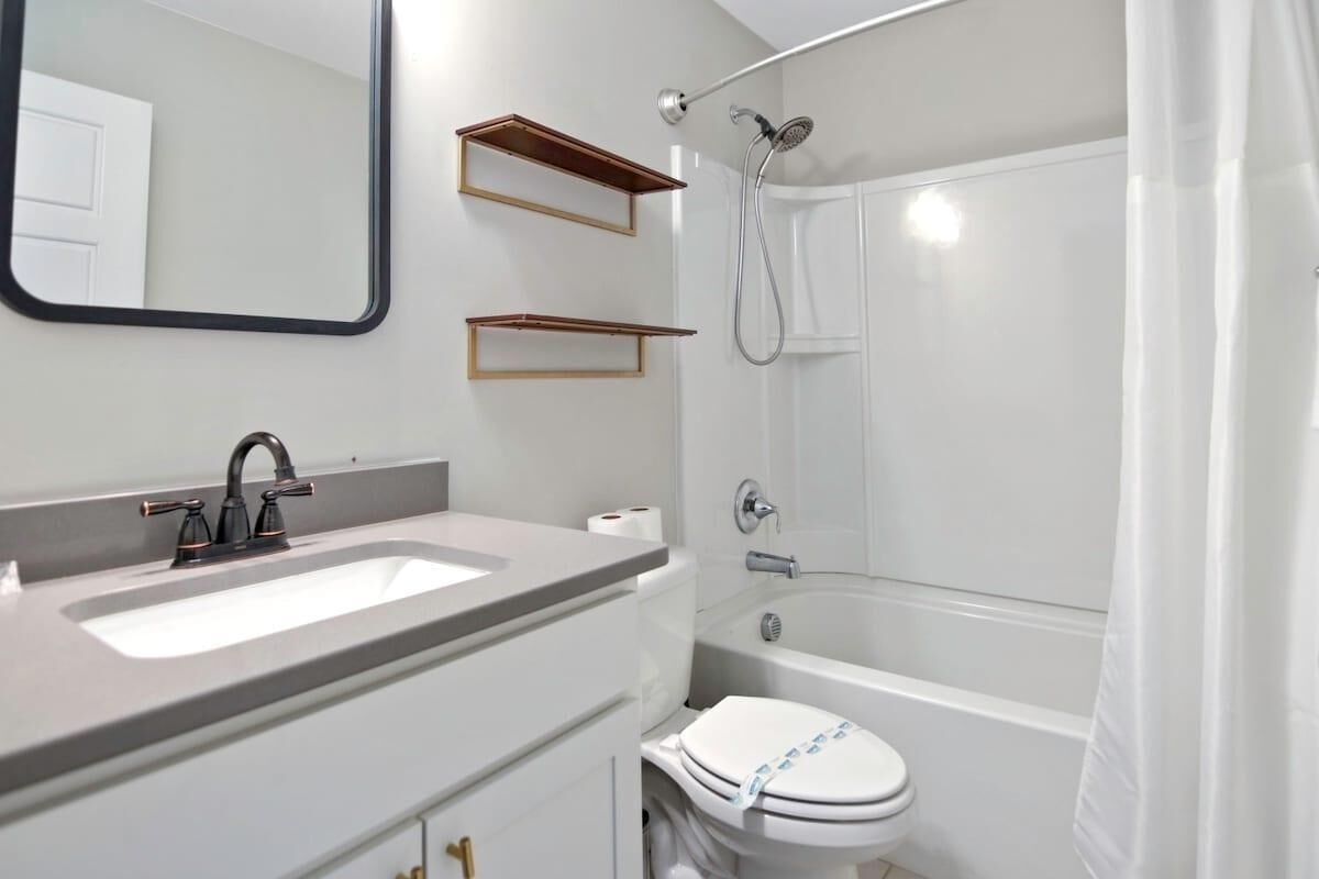 Un baño moderno con lavamanos, espejo y retrete blancos. Hay repisas de madera sobre el lavamanos y un cabezal de ducha sobre el retrete.
