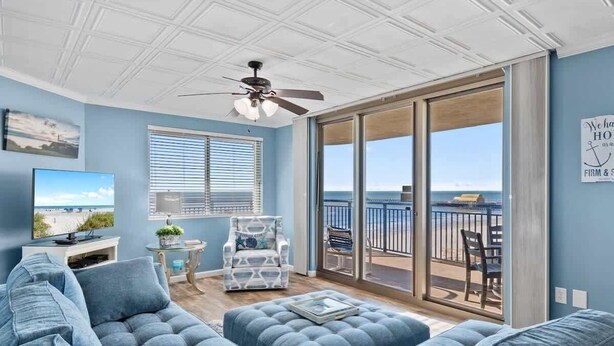 A living room with a large sofa, a patterned armchair, and a television. There is a balcony with a view of the ocean. - Oceanfront condo with balcony & resort pool (Myrtle Beach)
