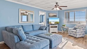A living room with a sectional sofa, a ceiling fan, a television, and a window with blinds. - Oceanfront condo with balcony & resort pool (Myrtle Beach)