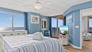 A bedroom with a large bed, a ceiling fan, a TV, and a view of the ocean. - Oceanfront condo with balcony & resort pool (Myrtle Beach)
