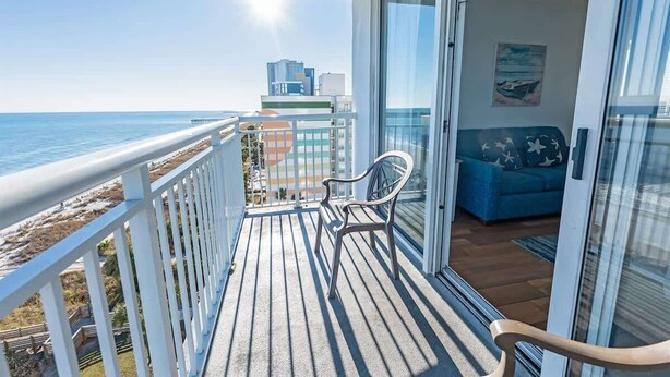 A balcony with a view of the ocean, a blue sofa, and a white chair. - Oceanfront condo with pool & hot tub (Myrtle Beach)