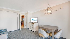 Hotellihuoneessa on ruokapöytä, kaksi tuolia, televisio, lipasto ja käytävä, joka johtaa keittiöön.