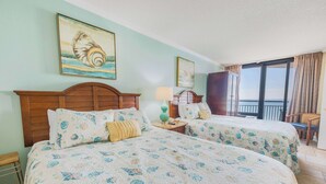 A hotel room with two beds, a wooden headboard, a nightstand, a lamp, and a view of the sea through a sliding door. - New Coastal Charm, Direct Oceanfront Views, Pools! (Myrtle Beach)